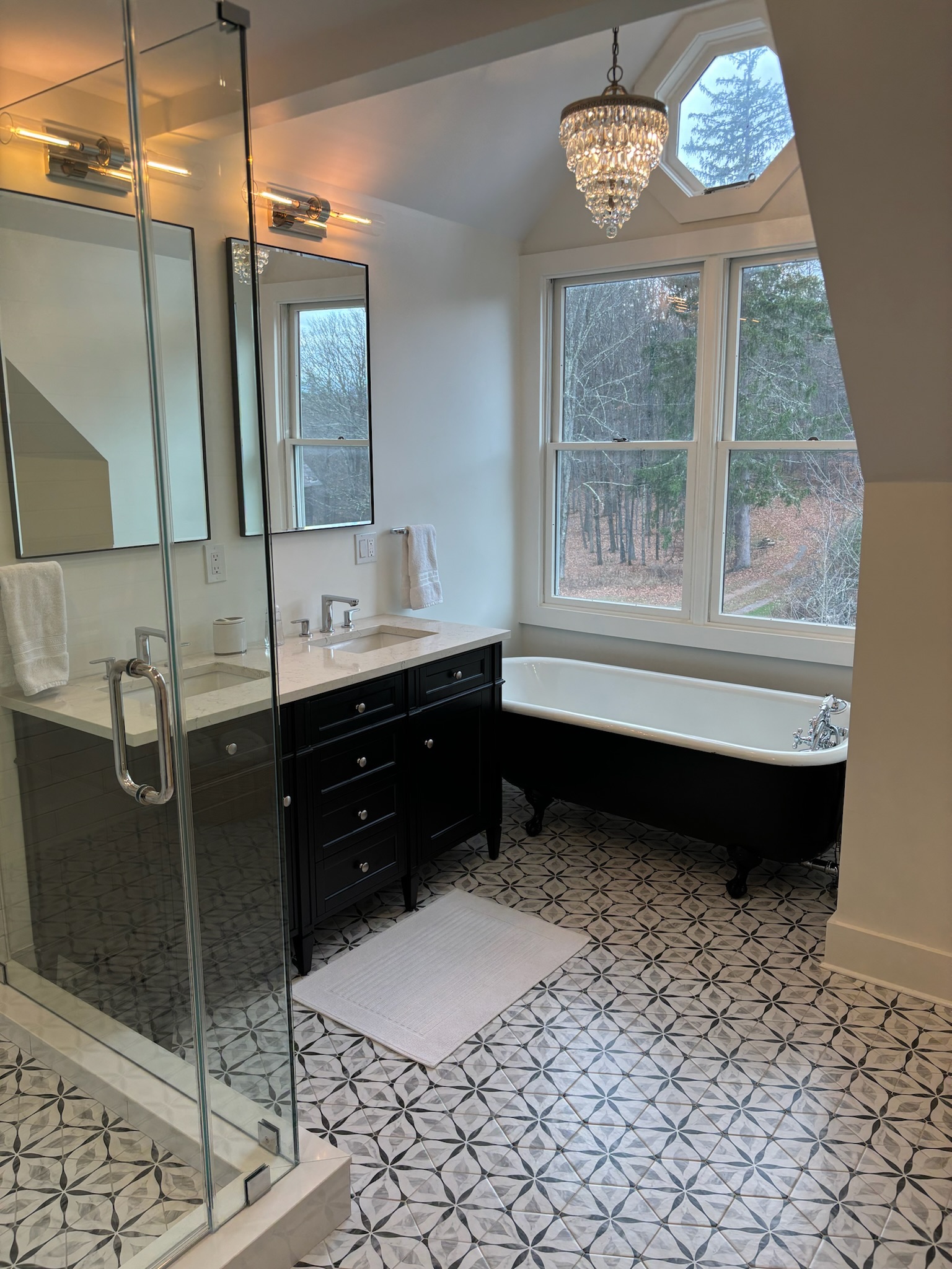 Master bathroom with black clawfoot soaking tub beneath windows overlooking forest, crystal chandelier, and geometric tile floor