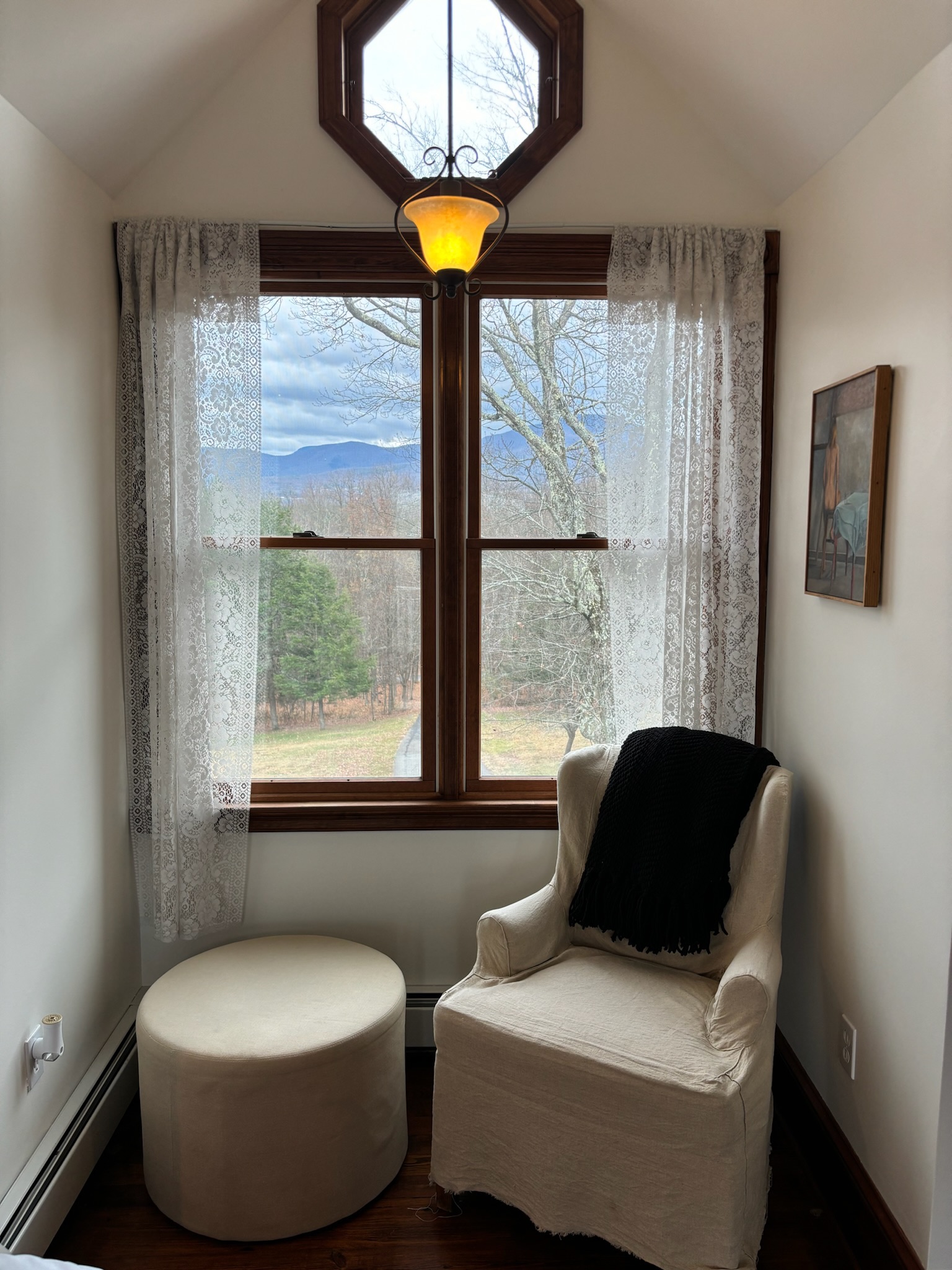 Master bedroom reading nook with wingback chair, round ottoman, octagonal transom window framing Catskill mountain views