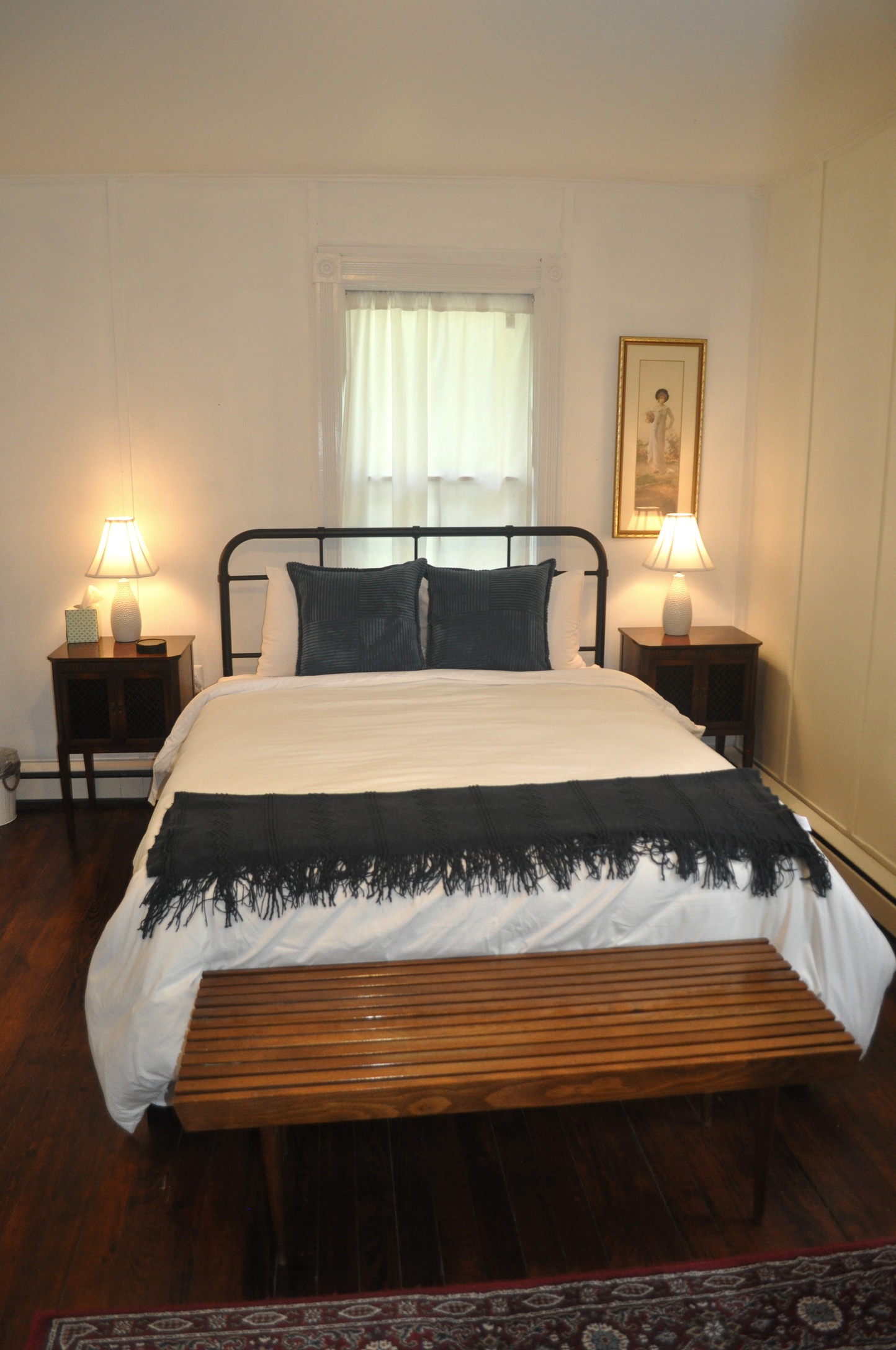 Bedroom two with double bed, iron headboard, white paneled millwork walls, lace curtain window, and hardwood floors