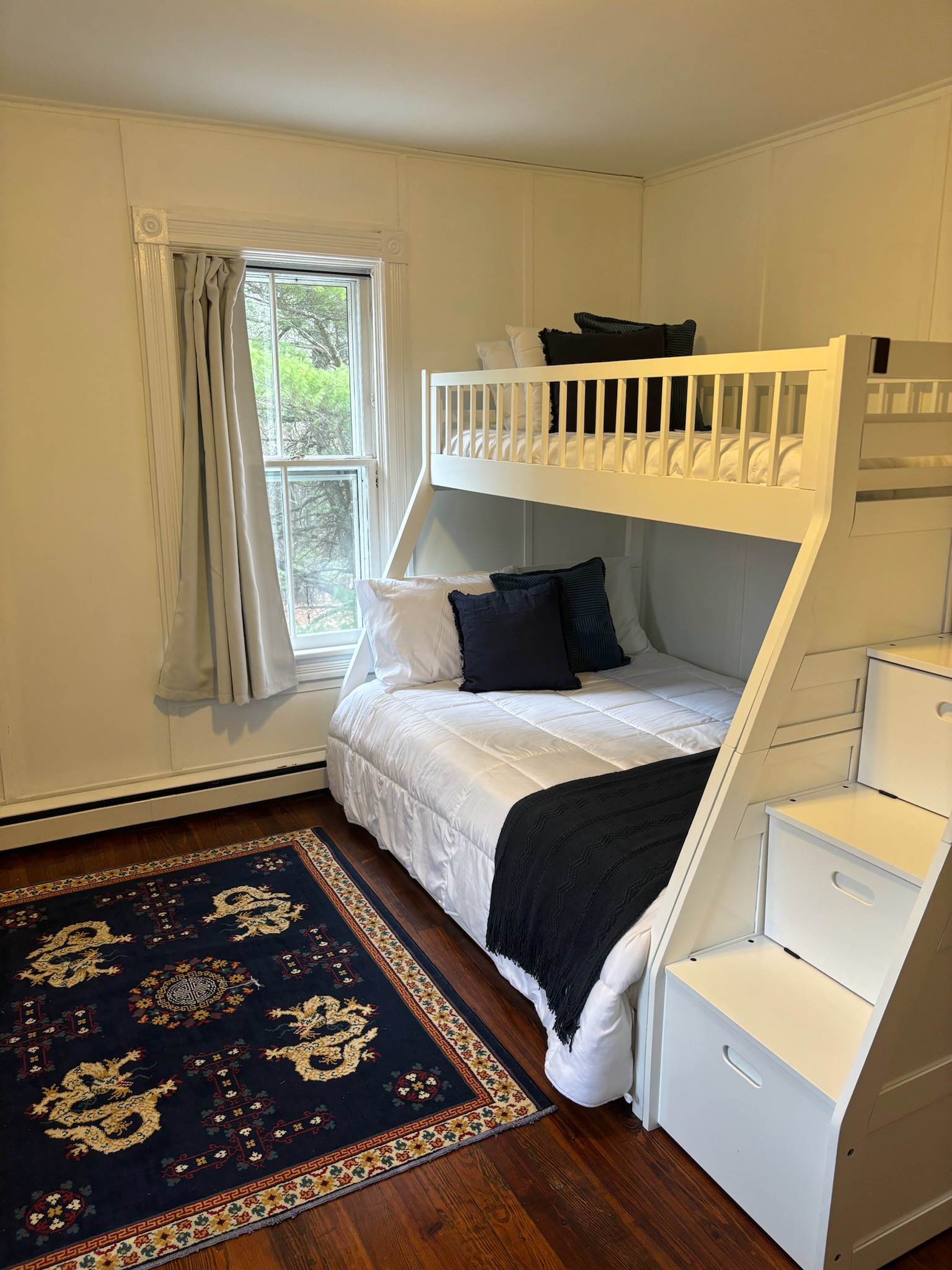 Bunk room with white twin-over-full bunk bed with built-in stair storage drawers, deep blue Chinese dragon rug, and navy armchair with ottoman