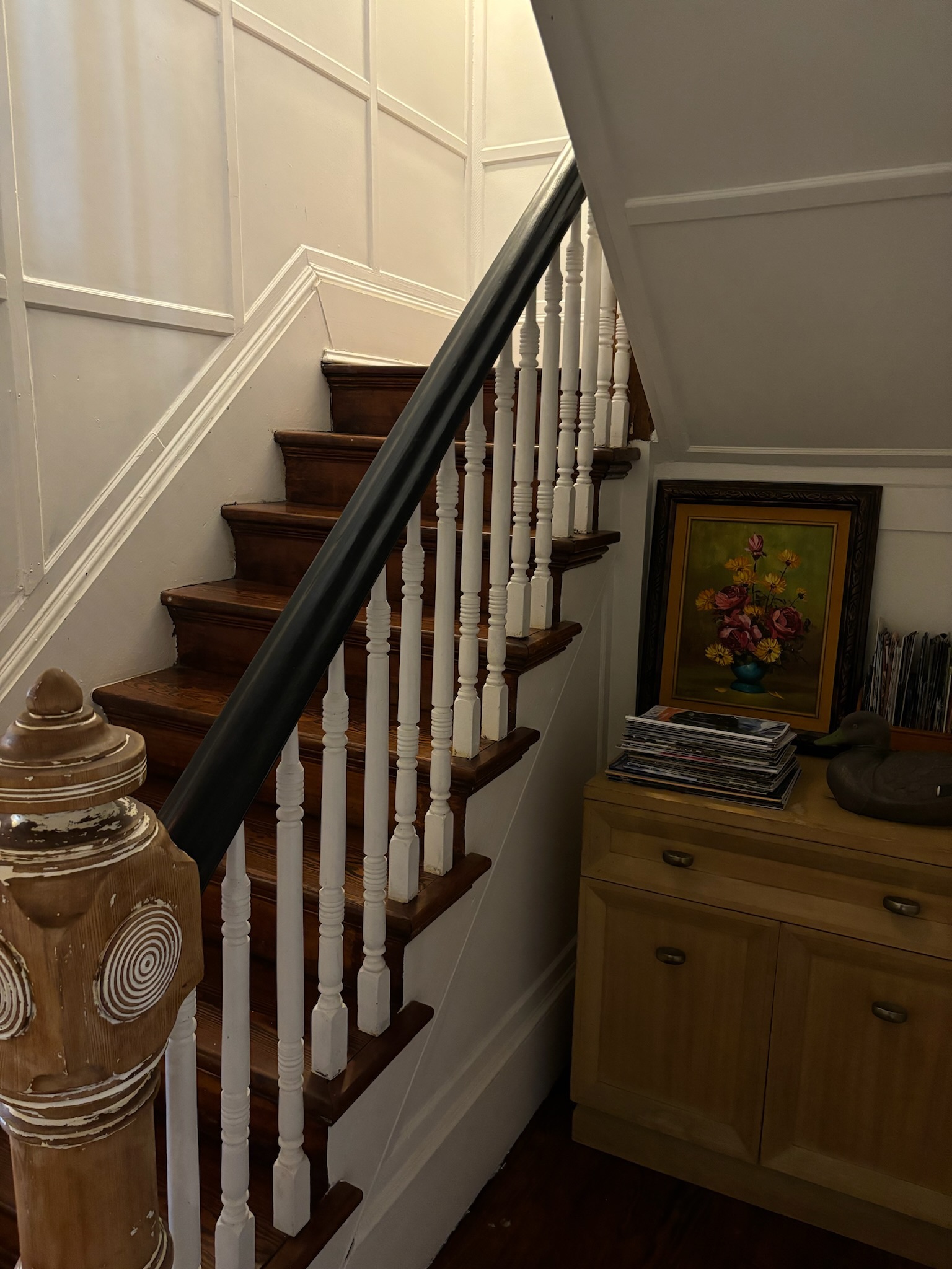 The original staircase with carved wood newel post, white turned balusters, black handrail, and framed oil painting on the wall beside it